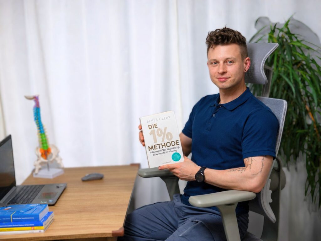 Schmerzcoach Ilja Panin aus Jena zeigt das Buch Die 1% Methode, um gesunde Gewohnheiten bei Rückenschmerzen aufzubauen.
